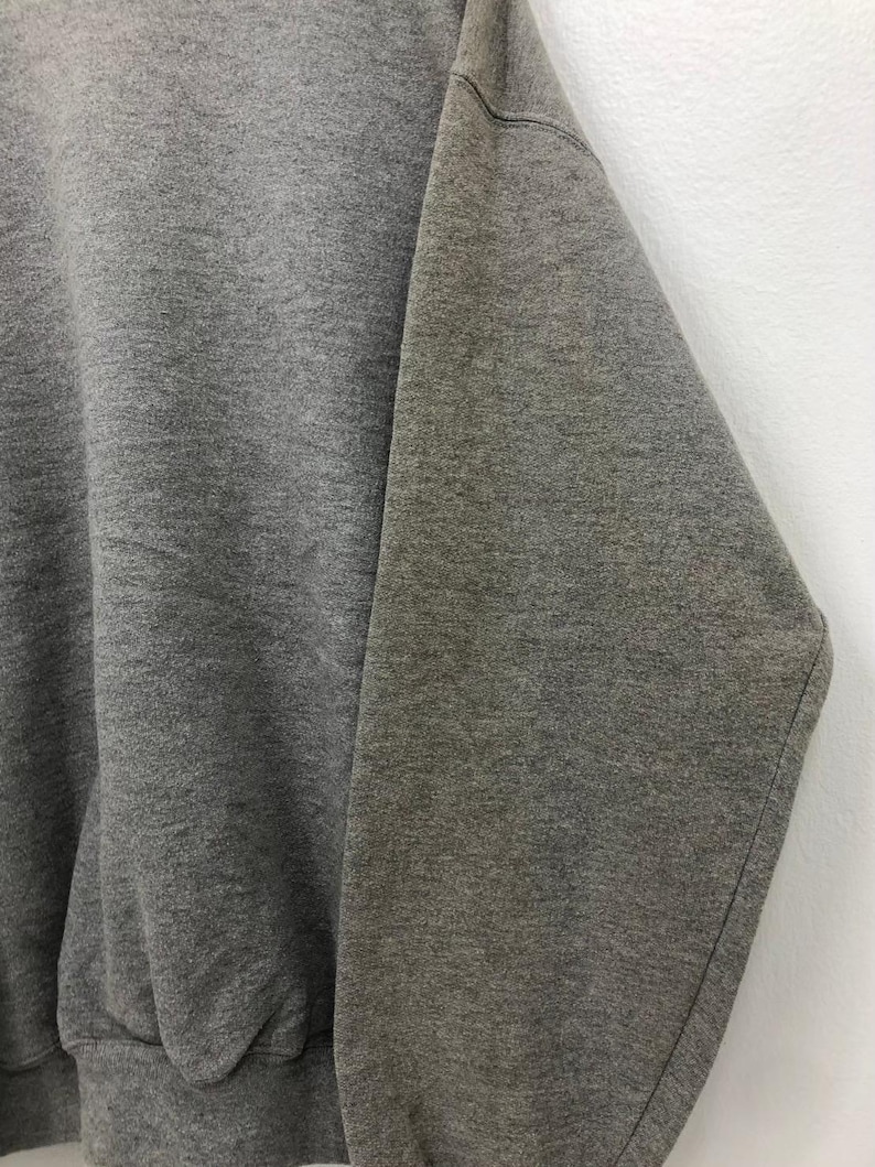 May include: Close-up of a gray sweatshirt. The fabric appears to be a soft, textured material. The image focuses on the sleeve and part of the body of the garment. The sweatshirt is a solid, medium gray color.