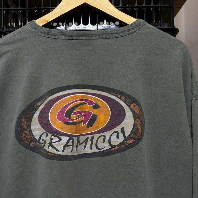 K&ouml;nnte beinhalten: Olivgr&uuml;nes Langarmshirt mit einem gro&szlig;en, ovalen Grafikdesign. Das Design zeigt ein stilisiertes "G" in Orange und Lila, umgeben von einem braunen und grauen Rand. Das Wort "GRAMICCI" ist darunter gedruckt.