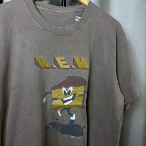 May include: Brown t-shirt featuring the band name "R.E.M." in yellow and brown block letters. Below the text is a cartoon image of a slice of cake with arms and legs. The shirt has a crew neck and short sleeves.