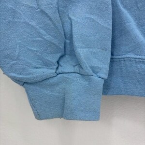 以下が含まれることがあります： 水色のスウェットシャツの袖のクローズアップ。リブ編みの袖口とギャザーの入った肘がカジュアルなスタイルを演出。生地は少し使い古されたように見えます。背景はプレーンな白い表面です。