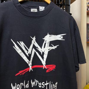 Pode incluir: Uma t-shirt preta de manga curta com um logótipo WWF grande e rabiscado a branco com um sublinhado rabiscado a vermelho. Abaixo do logótipo, o texto World Wrestling aparece a branco. Uma pequena etiqueta branca no decote exibe 22/28 A08 78.