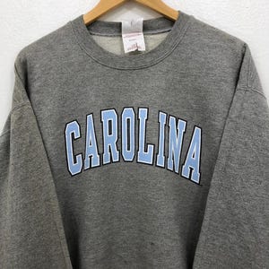 May include: A gray crewneck sweatshirt with the word "CAROLINA" in light blue block letters outlined in white across the chest. The sweatshirt is hanging on a wooden hanger.