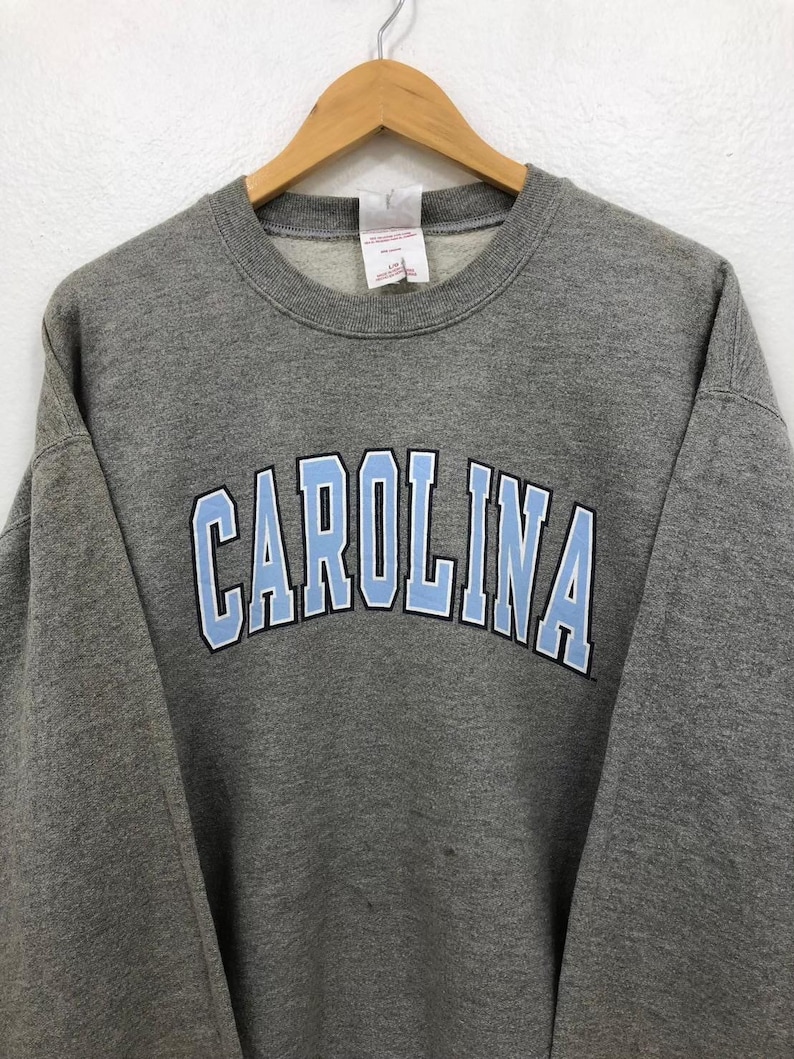 May include: A gray crewneck sweatshirt with the word "CAROLINA" in light blue block letters outlined in white across the chest. The sweatshirt is hanging on a wooden hanger.