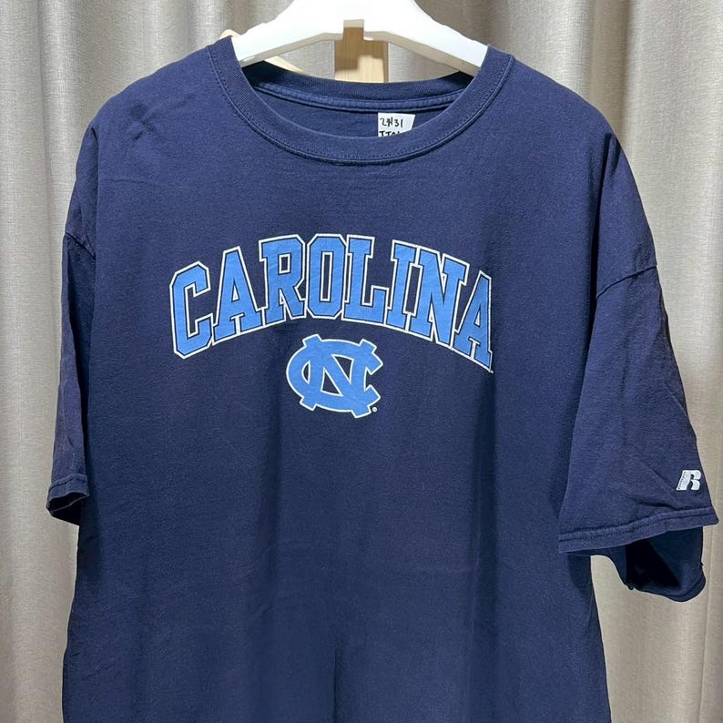 以下が含まれることがあります： 胸にライトブルーのアーチ状の「CAROLINA」の文字が入ったネイビーブルーのTシャツ。その下にライトブルーのロゴがあります。シャツは半袖で、クルーネックです。白いハンガーにかかっています。