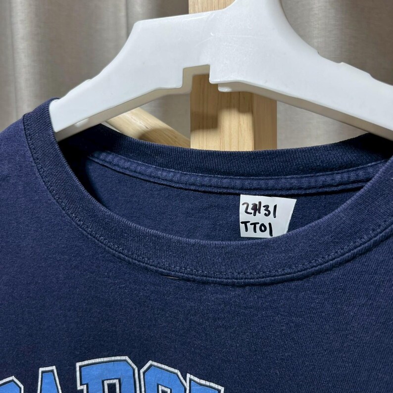 以下が含まれることがあります： 白いハンガーに掛けられた、ネイビーブルーのクルーネックTシャツ。シャツの襟のタグには「27/31」の数字と「TTO」の文字が書かれています。シャツの下部には白と青のデザインがあります。