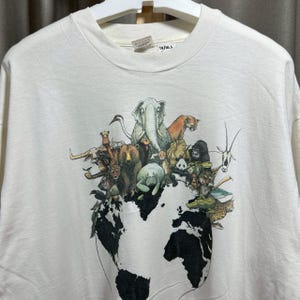 May include: Off-white t-shirt featuring a graphic of various animals, including an elephant, bear, and gorilla, clustered on top of a world map. The shirt has a crew neck and short sleeves. The tag reads "24/30.5".
