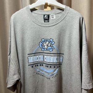May include: A gray t-shirt featuring the North Carolina Tar Heels logo. The logo includes the words "North Carolina Tar Heels" in a bold font, with a light blue and white color scheme. The shirt also has the text "Established 1789".