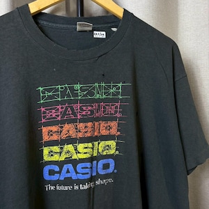 Könnte beinhalten: Schwarzes T-Shirt mit einem farbenfrohen Grafikdesign des Wortes "CASIO" in einem Raster. Der Text "The future is taking shape" ist unter dem Logo aufgedruckt. Das T-Shirt hängt an einem Holzkleiderbügel.
