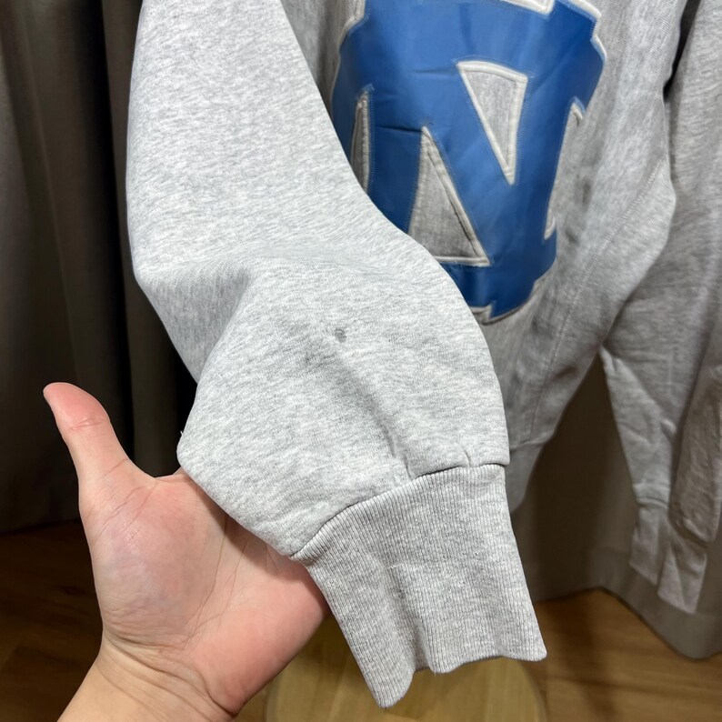 May include: A gray sweatshirt sleeve with a blue and white emblem. The emblem features a large letter "N" in blue, with a white outline. The sleeve is cuffed at the wrist. The sweatshirt appears to be vintage.