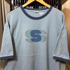 May include: Pale blue ringer t-shirt with a navy trim on the collar and sleeves. The shirt displays a layered "S" logo in blue and grey. The garment is suspended on a wooden hanger.