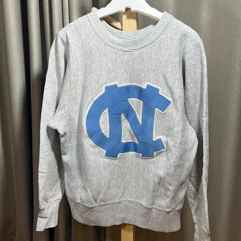 May include: A gray sweatshirt with a large light blue logo on the front. The logo is a stylized "NC" design. The sweatshirt has long sleeves and a crew neck. The garment is hanging on a white hanger.