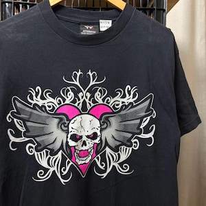 May include: A black short-sleeved t-shirt featuring a graphic print of a white skull with pink eyes and mouth, centred in a pink heart with grey wings and white swirling filigree patterns. The inner neck label displays the text Authentic World Wrestling.