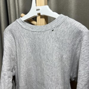 May include: A heather gray crew neck sweatshirt hanging on a white hanger. The sweatshirt has a small, dark-colored detail near the neckline. The fabric appears soft and comfortable, suitable for casual wear.