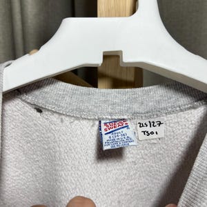 May include: Close-up of a gray sweatshirt with a crew neck, hanging on a white hanger. The tag reads "SOF SWEATS" and "ADULT S (34-36)". Additional text includes "MADE IN U.S.A." and "SEE REVERSE".