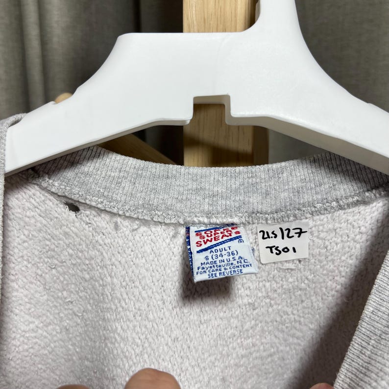 May include: Close-up of a gray sweatshirt with a crew neck, hanging on a white hanger. The tag reads "SOF SWEATS" and "ADULT S (34-36)". Additional text includes "MADE IN U.S.A." and "SEE REVERSE".