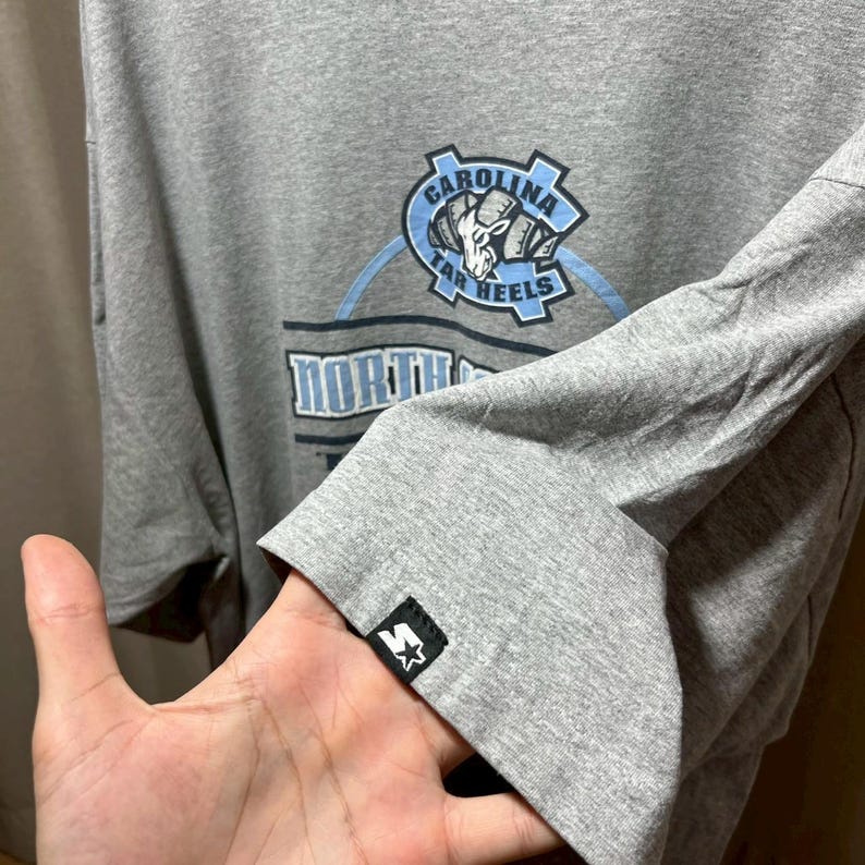 May include: Gray long-sleeve shirt featuring the Carolina Tar Heels logo in light blue and white. The shirt has the Starter logo on the sleeve cuff. The text "North" is visible on the shirt.