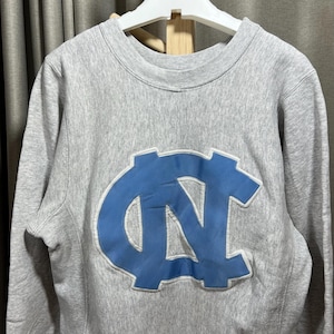 May include: A gray sweatshirt with a light blue logo on the front. The logo is a stylized interlocking "NC" design. The sweatshirt has a crew neck and long sleeves. The fabric appears to be a soft, comfortable material.