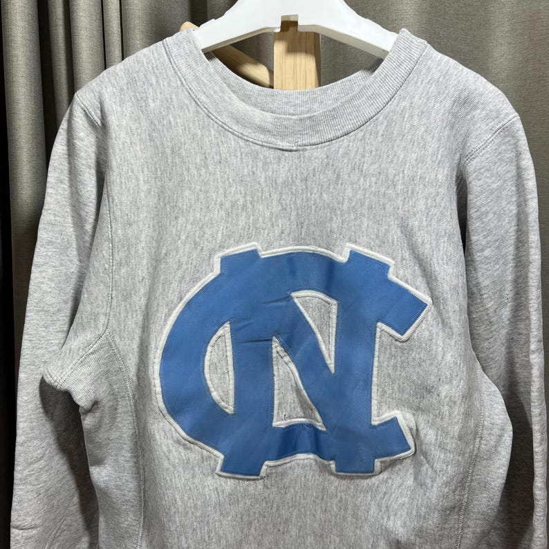 May include: A gray sweatshirt with a light blue logo on the front. The logo is a stylized interlocking "NC" design. The sweatshirt has a crew neck and long sleeves. The fabric appears to be a soft, comfortable material.