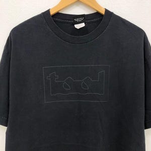 Może przedstawiać: Czarny t-shirt z szarym obrysem słowa "TOOL" w prostokątnym pudełku. Koszulka wisi na drewnianym wieszaku. T-shirt jest wykonany z miękkiego materiału.