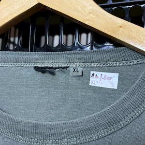 K&ouml;nnte beinhalten: Nahaufnahme eines grauen XL-T-Shirts mit Etikett. Das Shirt h&auml;ngt an einem Holzb&uuml;gel. Das Etikett hat unleserlichen Text und Zahlen. Das Shirt ist aus einem weichen Material.