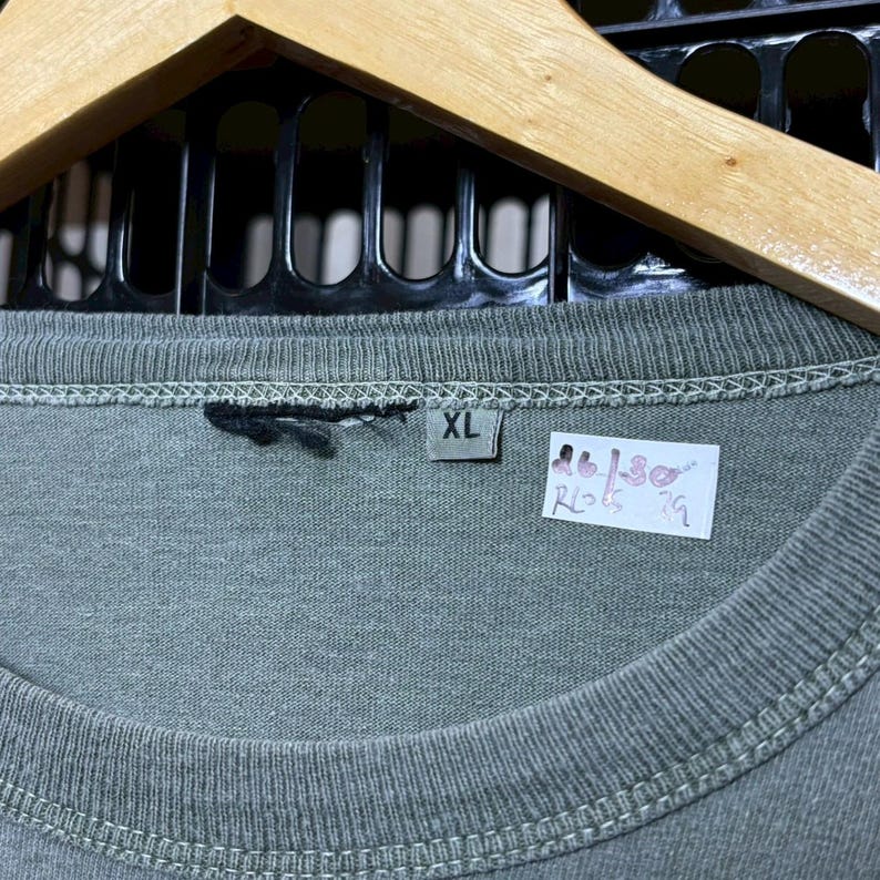 K&ouml;nnte beinhalten: Nahaufnahme eines grauen XL-T-Shirts mit Etikett. Das Shirt h&auml;ngt an einem Holzb&uuml;gel. Das Etikett hat unleserlichen Text und Zahlen. Das Shirt ist aus einem weichen Material.