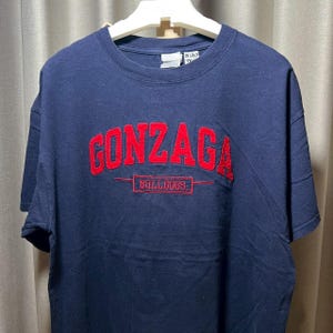 May include: Navy blue t-shirt with the word "GONZAGA" in red, embroidered across the chest. Below the word "GONZAGA" is the word "BULLDOGS" in red, inside a red rectangle. The shirt is hanging on a white hanger.