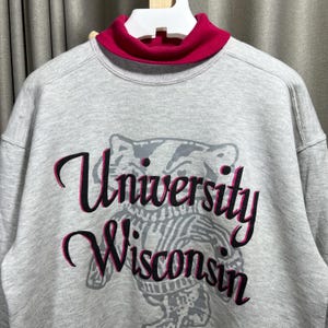 May include: A gray sweatshirt with a red turtleneck collar. The front features a graphic of a badger and the words "University Wisconsin" in black and pink script. The sweatshirt is a casual style.