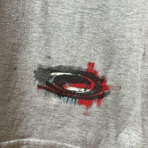 May include: Close-up of a gray t-shirt featuring a graphic design. The design is a stylized, abstract shape in black, white, and red, with a splatter effect. The shirt appears to be made of a soft, worn fabric.