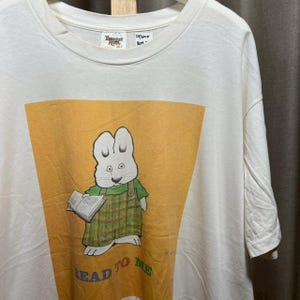 Puede incluir: Camiseta blanca con un conejo de dibujos animados sosteniendo un libro, con un peto verde con estampado de cuadros. El texto "READ TO ME" está impreso en la parte inferior del diseño. La camiseta tiene cuello redondo y mangas cortas.