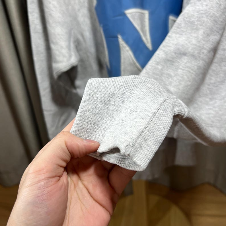 May include: A gray sweatshirt with a large blue letter "N" on the front. The sleeve cuff is visible, showing the ribbed knit fabric. The sweatshirt appears to be made of a soft, comfortable material.