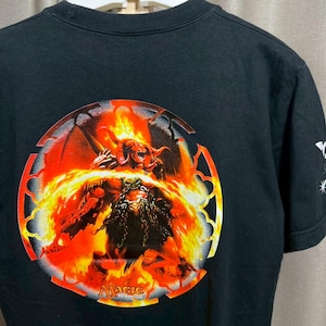 Può includere: T-shirt nera con una grafica circolare raffigurante una figura demoniaca in fiamme. Il design include il testo "Magic: The Gathering" in basso. La maglietta è realizzata in materiale morbido.