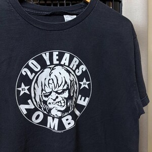 May include: A black short-sleeve t-shirt featuring a white circular graphic on the chest. The graphic displays a stylized zombie face in the center, surrounded by the text 20 YEARS ZOMBIE, with the numbers 85 and 05 placed inside small stars.