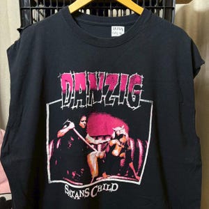 May include: Black sleeveless t-shirt with a pink and white graphic. The graphic features the band name "DANZIG" and the words "SATANS CHILD" with an illustration of two figures. The shirt is hanging on a wooden hanger.