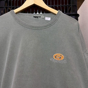 K&ouml;nnte beinhalten: Verblasstes olivgr&uuml;nes T-Shirt mit Rundhalsausschnitt und kurzen &Auml;rmeln. Das Gramicci-Logo in Orange und Wei&szlig; ist auf der Vorderseite abgebildet. Das T-Shirt h&auml;ngt an einem Holzb&uuml;gel.