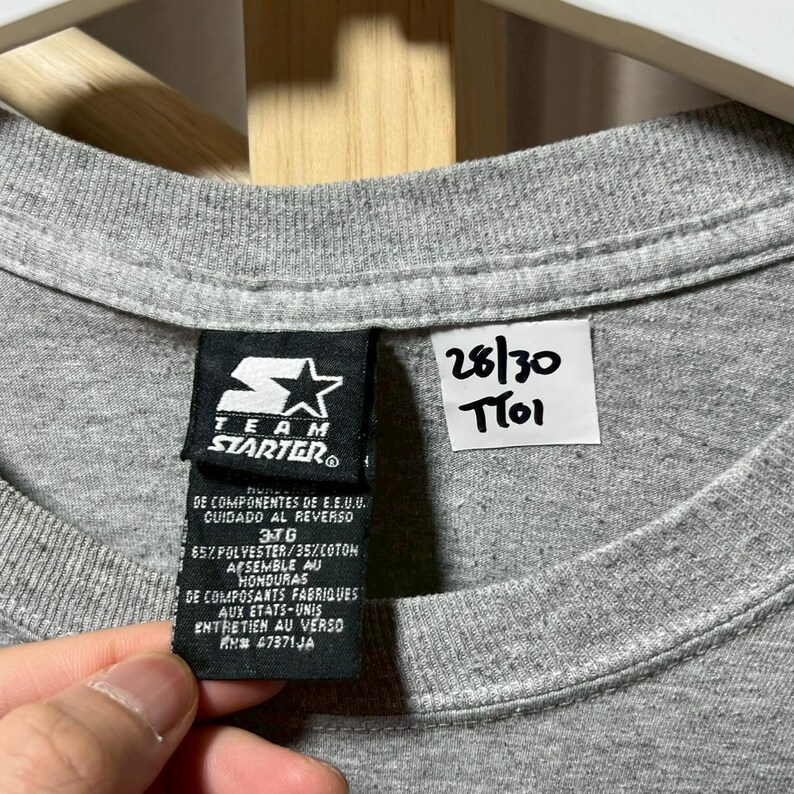 May include: Close-up of a gray t-shirt with a Starter brand tag. The tag includes text in English and Spanish, with details about the fabric composition and care instructions. A small white tag with the numbers "28/30" and "T101" is also visible.