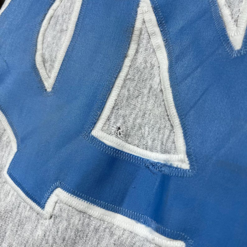May include: Close-up of a gray sweatshirt with a large, light blue letter "Y" applique. The letter is outlined in white and stitched onto the gray fabric. The image shows the texture of the fabric and stitching.