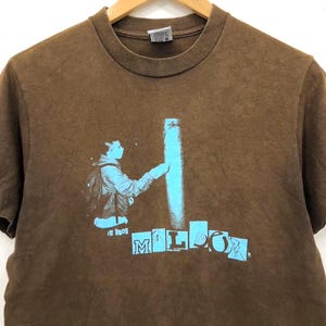 May include: Brown t-shirt with a blue graphic of a person spraying paint on a wall, and the word "MILLOR" in blue block letters. The shirt is displayed on a wooden hanger.