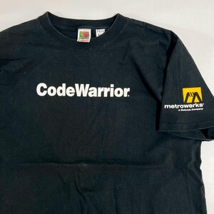 Puede incluir: Camiseta negra con la palabra "CodeWarrior" en letras blancas en el pecho. La manga derecha presenta el logotipo de "metrowerks" en amarillo y negro. Una etiqueta vintage de Fruit of the Loom es visible en el cuello.