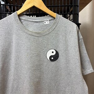 Puede incluir: Una camiseta gris jaspeado de manga corta con un pequeño símbolo de yin-yang en blanco y negro estampado en el lado izquierdo del pecho. Una etiqueta rectangular blanca con el texto negro 225/29 RD 2 está unida al interior del cuello redondo.