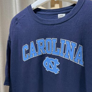 以下が含まれることがあります： 胸に白と水色で「CAROLINA」の文字がアーチ状に描かれたネイビーブルーのTシャツ。その下に水色のロゴがあります。白いハンガーにかかっています。
