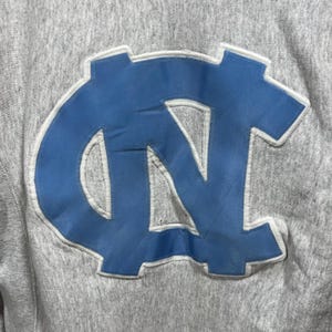 May include: Close-up of a gray sweatshirt featuring a large, light blue and white embroidered logo. The logo is a stylized "NC" design, with a white outline. The sweatshirt has a textured appearance.