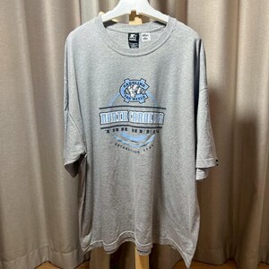 May include: A gray t-shirt with a crew neck and short sleeves. The shirt features a blue and white graphic that reads "North Carolina Tar Heels" with a logo and the text "Established 1789". The shirt is hanging on a white hanger.