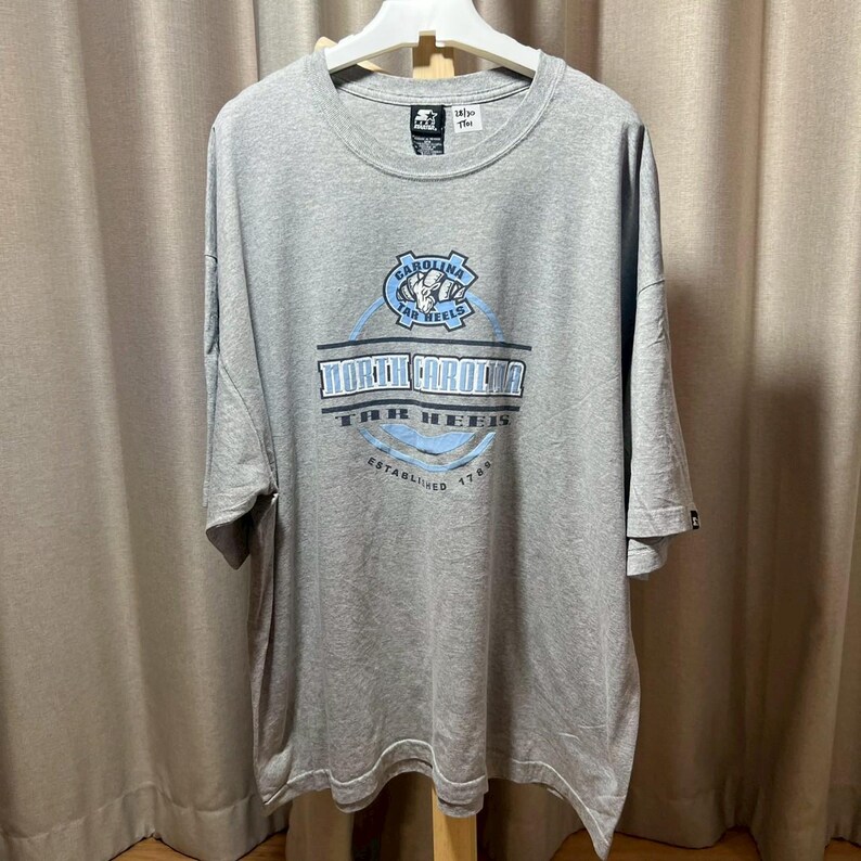 May include: A gray t-shirt with a crew neck and short sleeves. The shirt features a blue and white graphic that reads "North Carolina Tar Heels" with a logo and the text "Established 1789". The shirt is hanging on a white hanger.