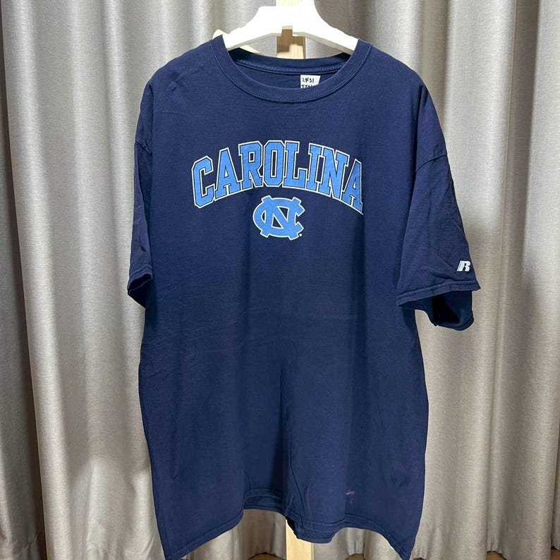 以下が含まれることがあります： 胸にライトブルーの「CAROLINA」の文字がアーチ状に描かれたネイビーブルーのTシャツ。その下にライトブルーのロゴがあります。このシャツは半袖で、クルーネックです。
