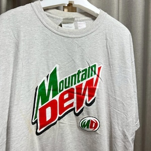 以下が含まれることがあります： 緑、赤、白のMountain Dewロゴが入ったライトグレーのTシャツ。Tシャツはクルーネックと半袖です。小さなMDロゴも表示されています。