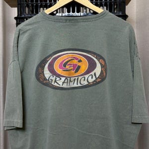 K&ouml;nnte beinhalten: Olivgr&uuml;nes T-Shirt mit einem gro&szlig;en, ovalen Grafikdesign auf dem R&uuml;cken. Die Grafik zeigt ein stilisiertes "G" in einem Kreis mit orangefarbenen, lila und wei&szlig;en Akzenten und das Wort "GRAMICCI" darunter.