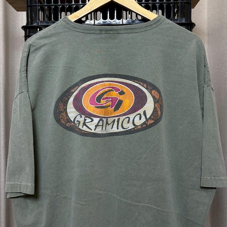 K&ouml;nnte beinhalten: Olivgr&uuml;nes T-Shirt mit einem gro&szlig;en, ovalen Grafikdesign auf dem R&uuml;cken. Die Grafik zeigt ein stilisiertes "G" in einem Kreis mit orangefarbenen, lila und wei&szlig;en Akzenten und das Wort "GRAMICCI" darunter.