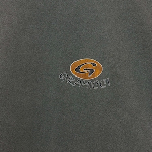 K&ouml;nnte beinhalten: Dunkelgr&uuml;nes Kleidungsst&uuml;ck mit goldenem und braunem Gramicci-Logo. Das Logo zeigt ein stilisiertes "G" in einem kreisf&ouml;rmigen Emblem, mit dem Markennamen darunter. Der Stoff scheint ein weiches, l&auml;ssiges Material zu sein.