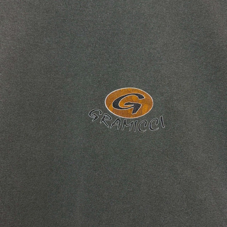 K&ouml;nnte beinhalten: Dunkelgr&uuml;nes Kleidungsst&uuml;ck mit goldenem und braunem Gramicci-Logo. Das Logo zeigt ein stilisiertes "G" in einem kreisf&ouml;rmigen Emblem, mit dem Markennamen darunter. Der Stoff scheint ein weiches, l&auml;ssiges Material zu sein.