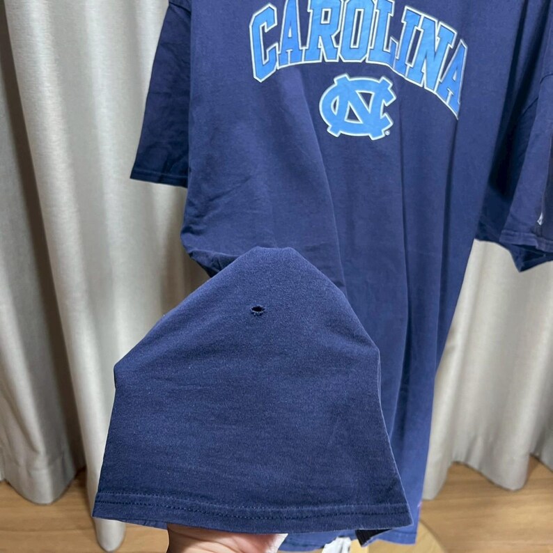以下が含まれることがあります： ライトブルーの文字で「CAROLINA」とライトブルーのロゴが入ったネイビーブルーのTシャツ。生地に小さな穴があります。ヴィンテージアイテムです。